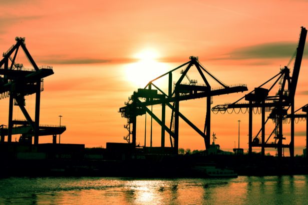 Image of a shipyard with three cranes with an orange sunset in the background.